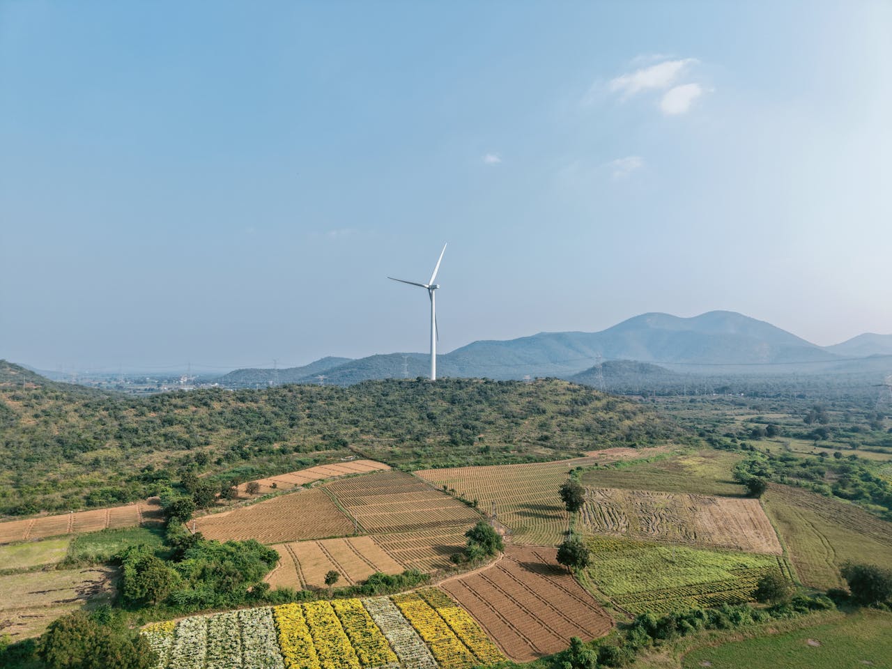 Aerial view of a wind turbine amidst rolling hills and farmland, under a clear blue sky.